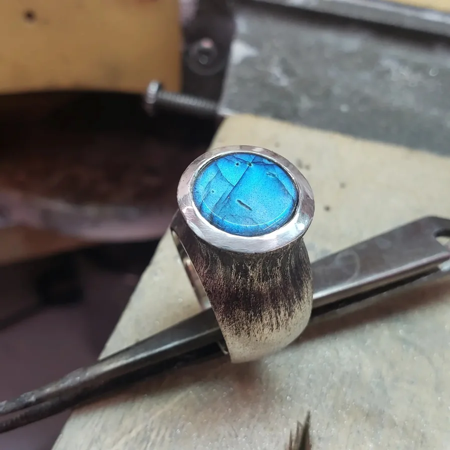 At the workbench, a bright blue labradorite held in its silver setting with a steel tool alongside.
