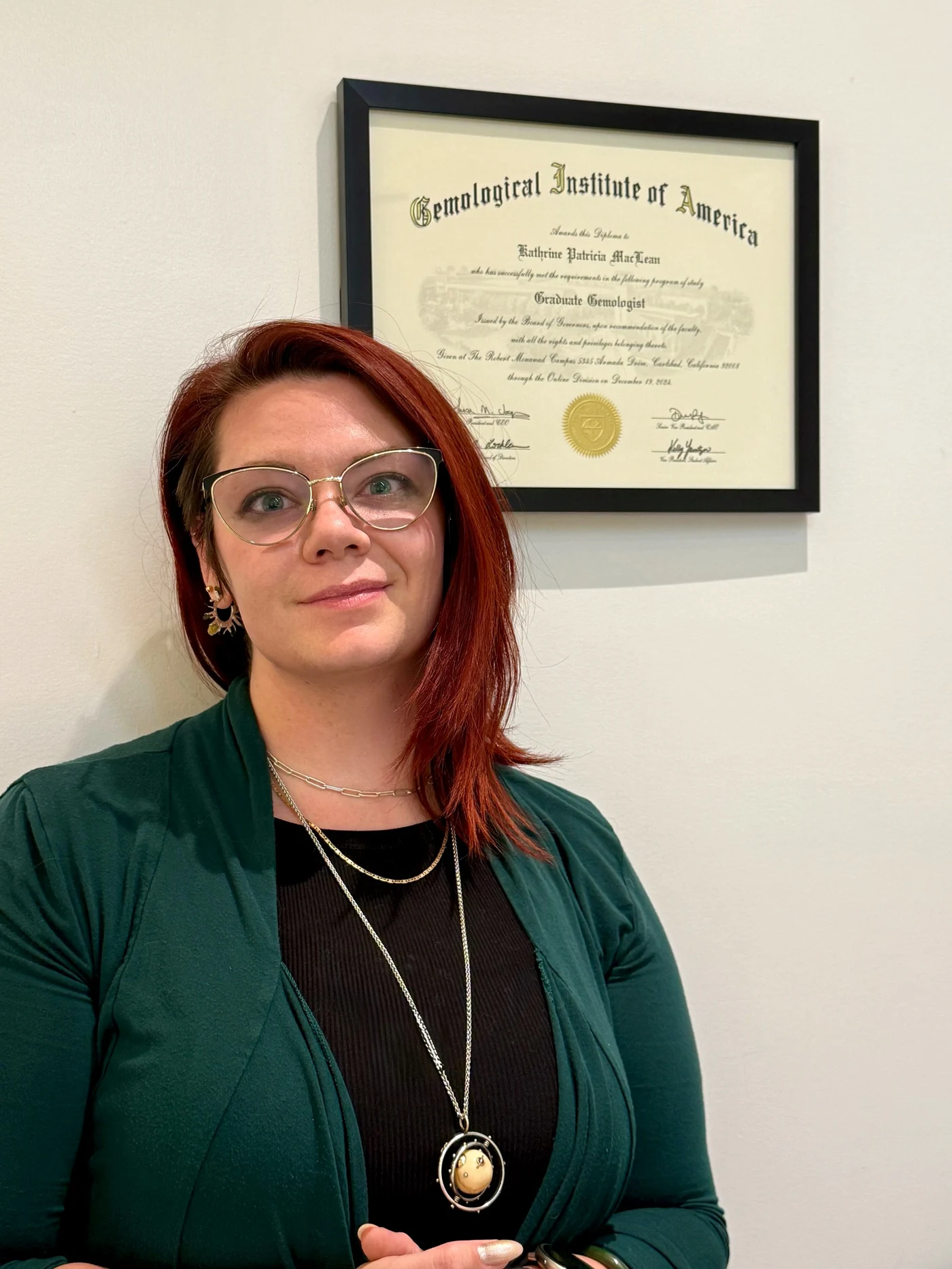 Kat MacLean standing in front of her framed Gemological Institute of America Graduate Gemologist diploma.