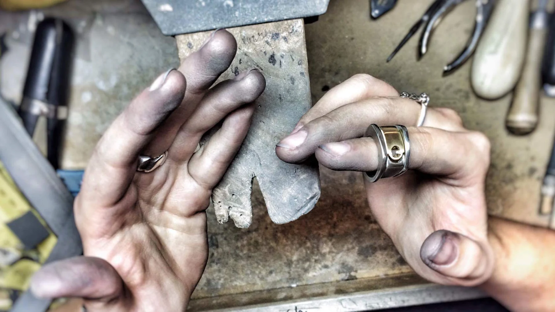 Kat's hands at her workbench, holding a metal template against a bench pin with pliers and burnishers around them.