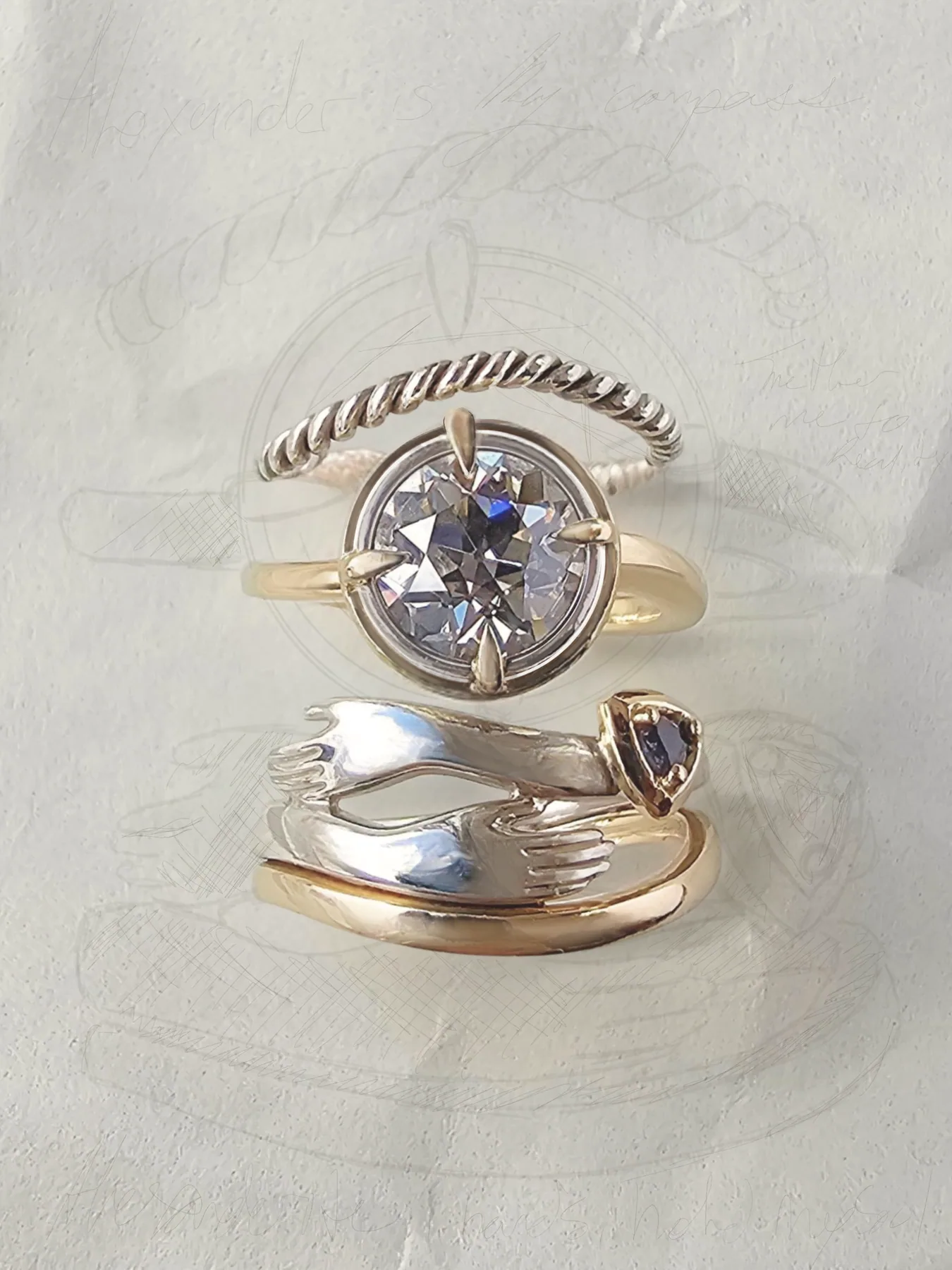 Three of Kat's own rings stacked on white paper over her pencil drawing of them: a twisted silver band, a round diamond in a gold compass setting, and two silver hands holding a small purple stone.