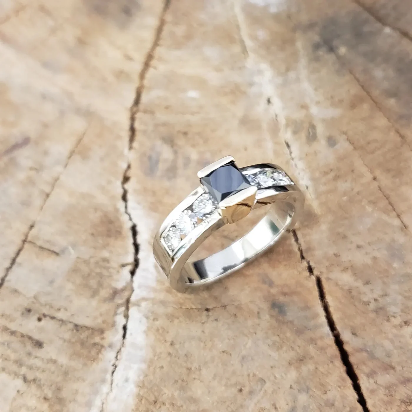 A white gold ring set with a princess cut black diamond flanked by channel set white diamonds, photographed on weathered pale wood.