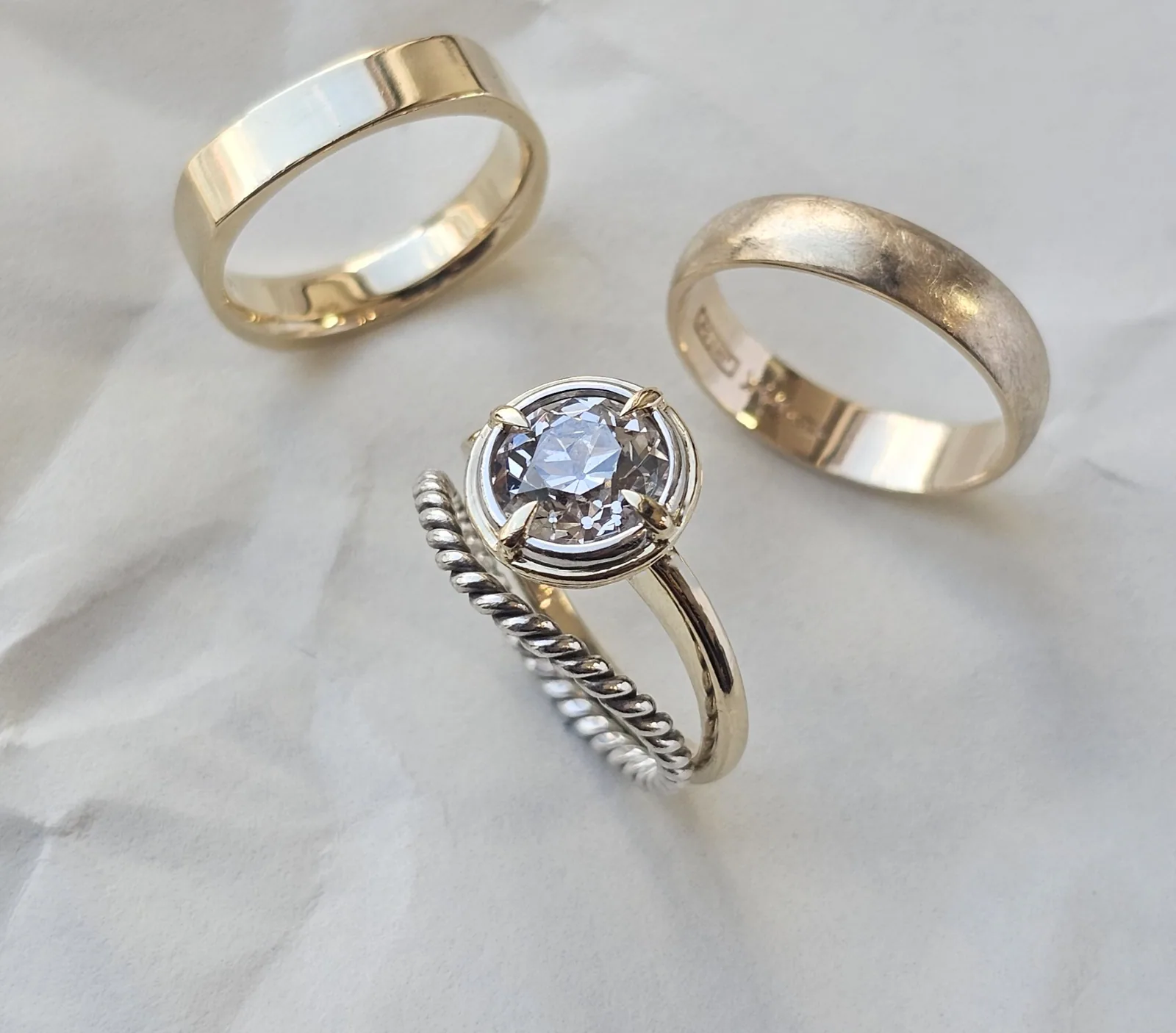 Two yellow gold wedding bands beside a round diamond held by four gold claws in a compass style setting on a twisted silver band, photographed on crumpled white paper.