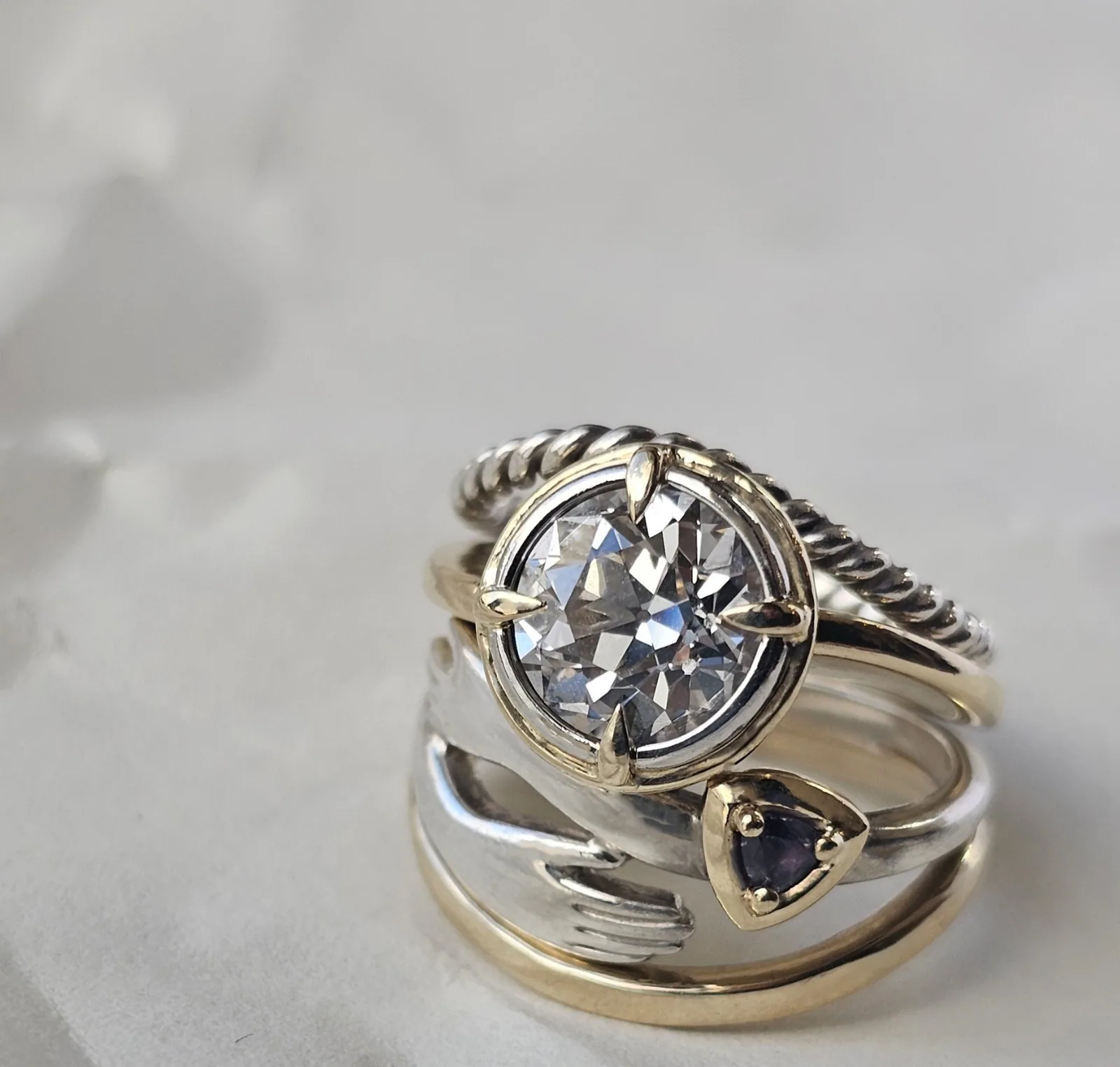 Three rings stacked at an angle on crumpled white paper: a twisted silver band, a round diamond in a gold compass setting, and a ring of two clasped silver hands holding a trillion cut purple stone on a gold band.