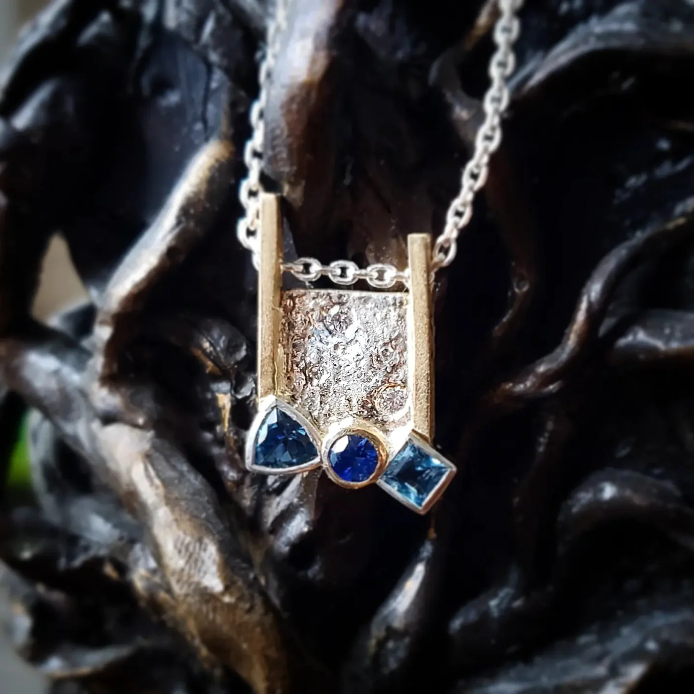A textured silver pendant flanked by two gold posts and set with three blue stones, hanging against dark twisted driftwood.