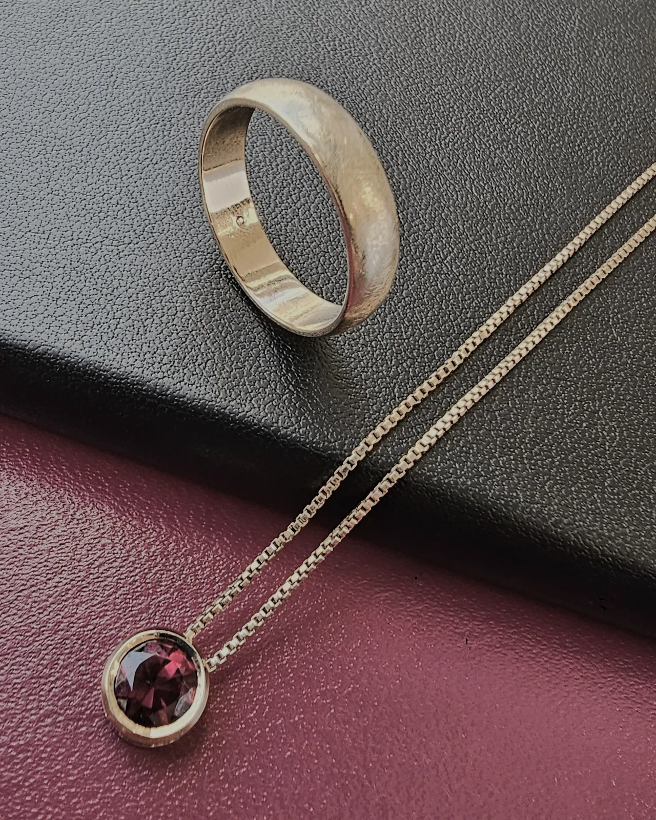 A round deep red stone in a plain bezel pendant on a fine box chain, beside a domed gold band, on black and burgundy leather.