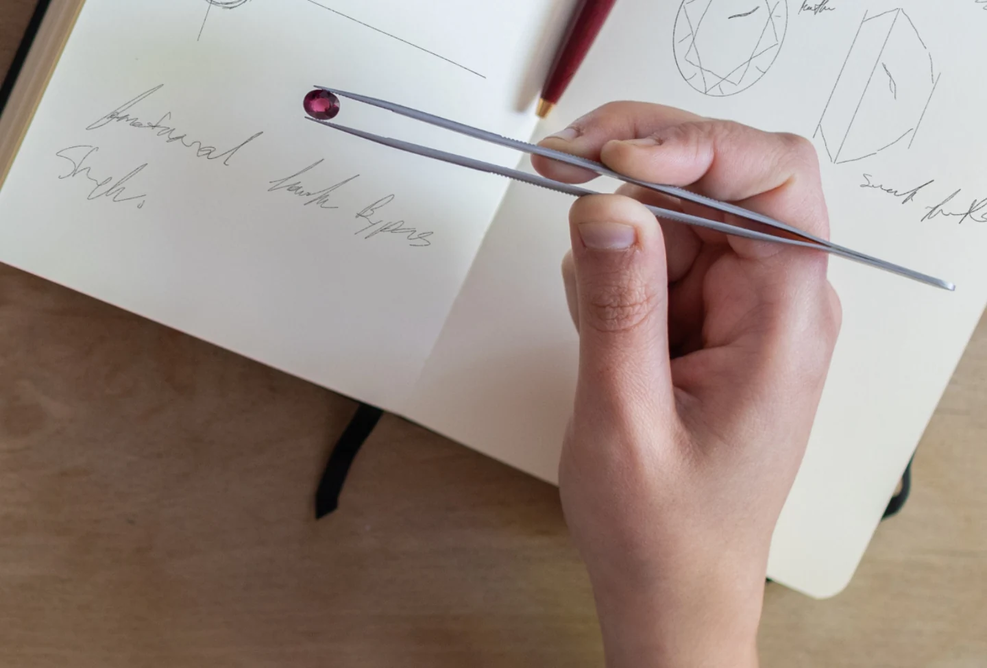 A hand holding tweezers with a red garnet, over an open Katalyst notebook showing handwritten stone measurements and ring sketches.